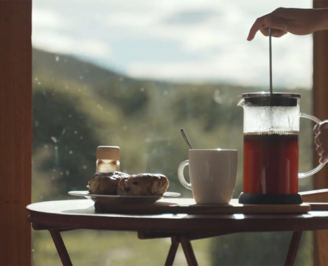 Gezellig koffiemoment met cafetière, mok en scones bij een raam met groen heuvelland onder een lichte bewolkte lucht.