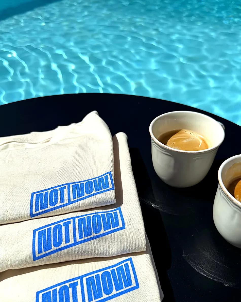 Drie opgevouwen beige NOT NOW t-shirts met blauw logo naast twee kopjes espresso op een tafel bij een zwembad.