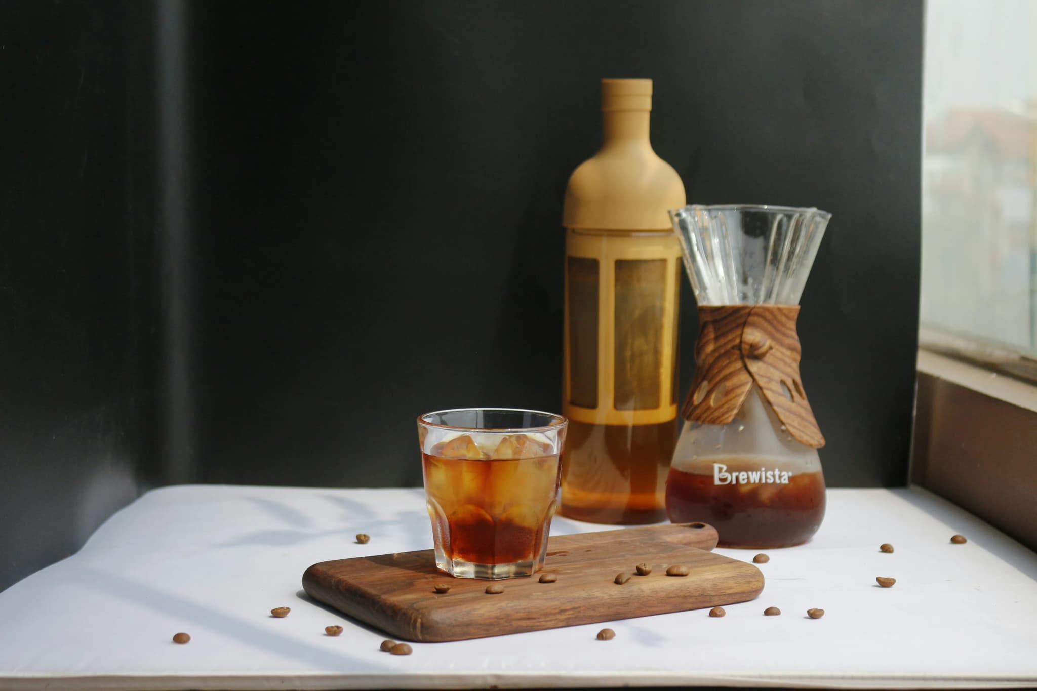 Close-up of a glass of cold brew coffee with ice on a wooden board, surrounded by coffee beans and coffee accessories on a dark background.