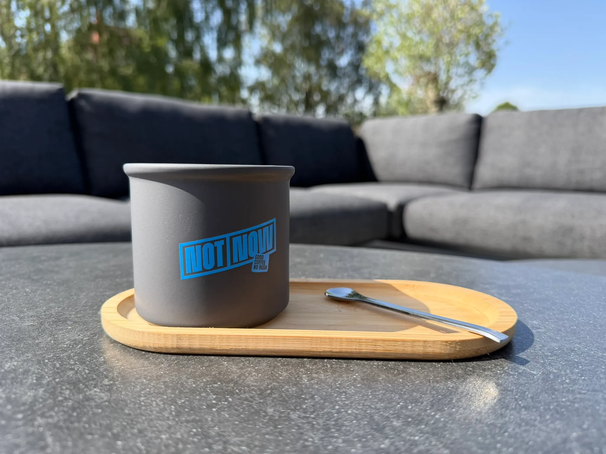 Donkergrijze keramische beker met blauw opschrift “NOT NOW” op een bamboe dienblad, naast een koffielepel.