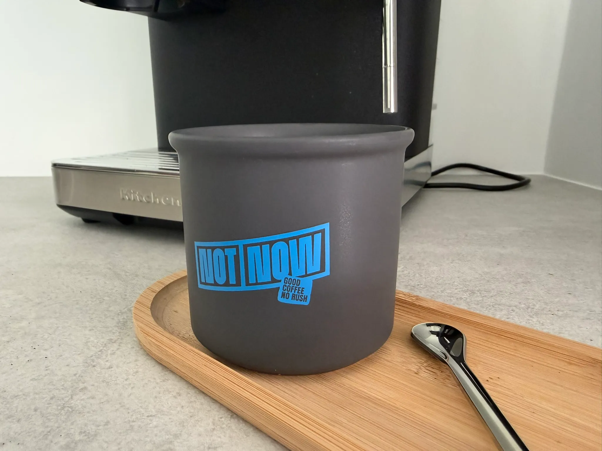 Donkergrijze koffiebeker met blauw-zwart ‘NOT NOW’-logo op een bamboe dienblad naast een koffiemachine.