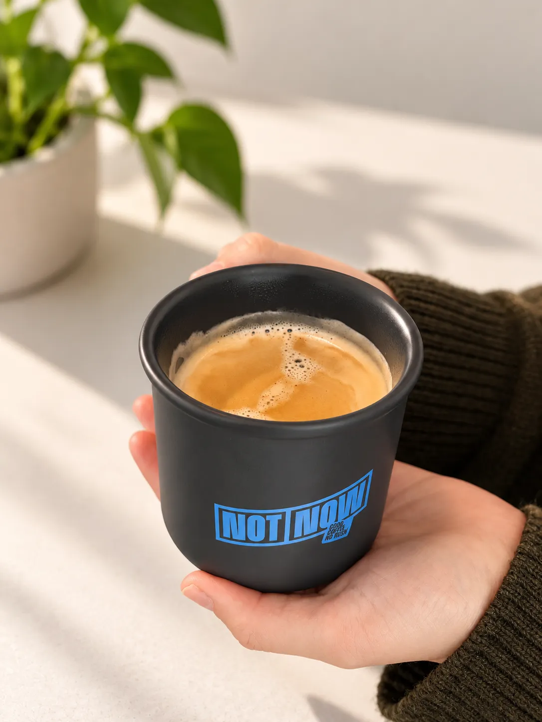 Handen in een donkergroene trui houden een zwarte koffiemok met blauw Not Now-logo in warm zonlicht vast.