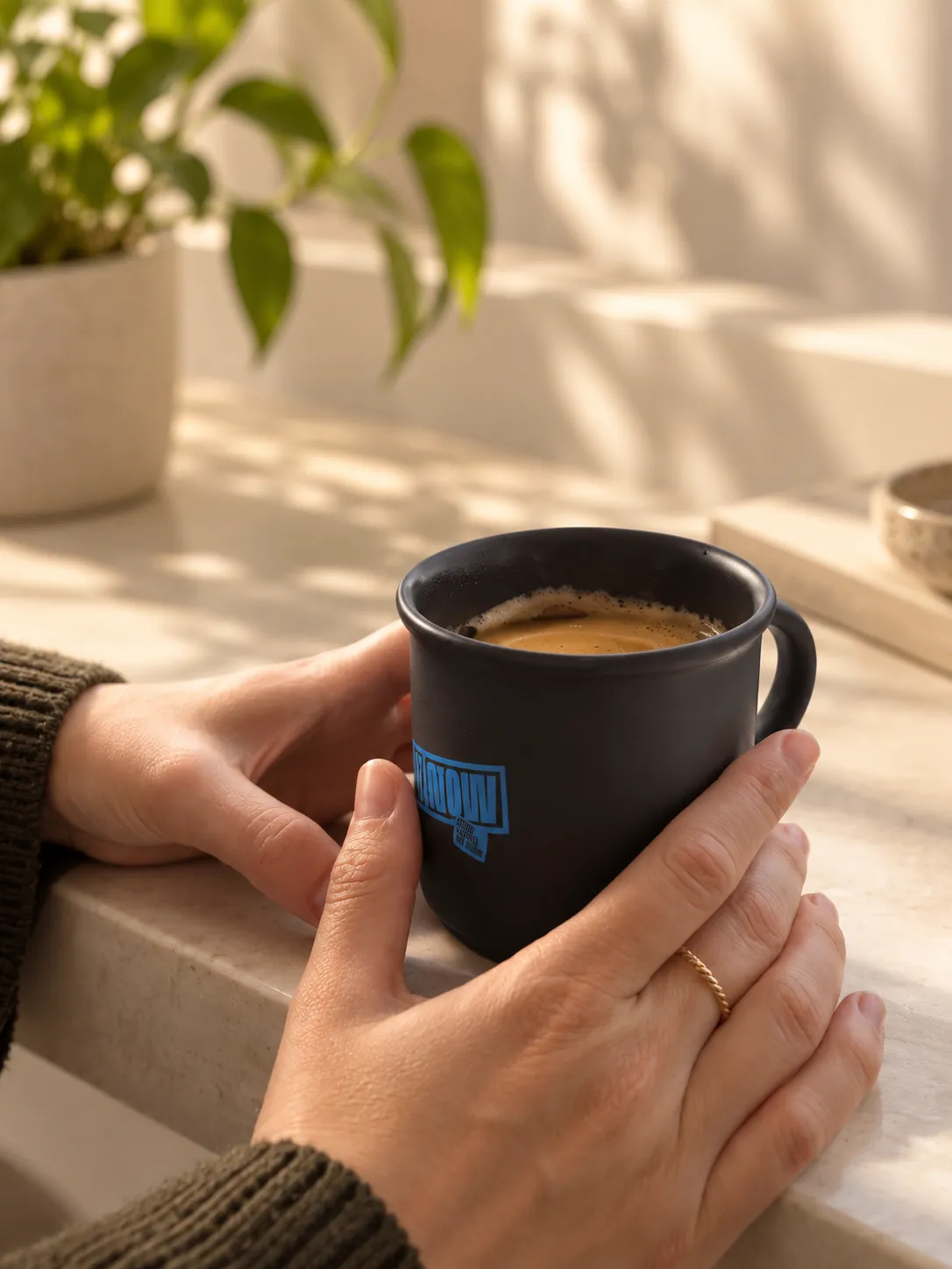 Handen houden een matzwarte koffiemok met blauwe opdruk en koffie vast op een vensterbank naast een kamerplant.